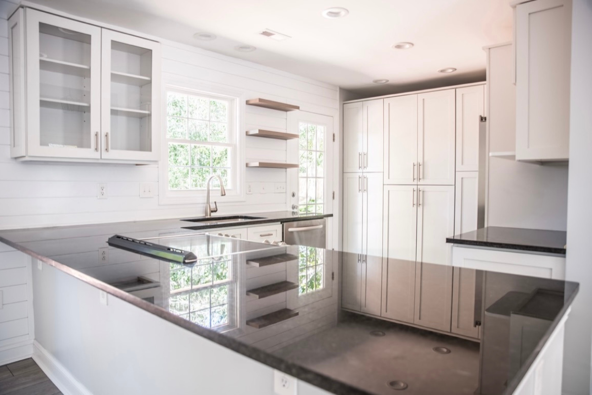 Modern white kitchen with glass-front cabinets, dark countertops, and open shelving near window
